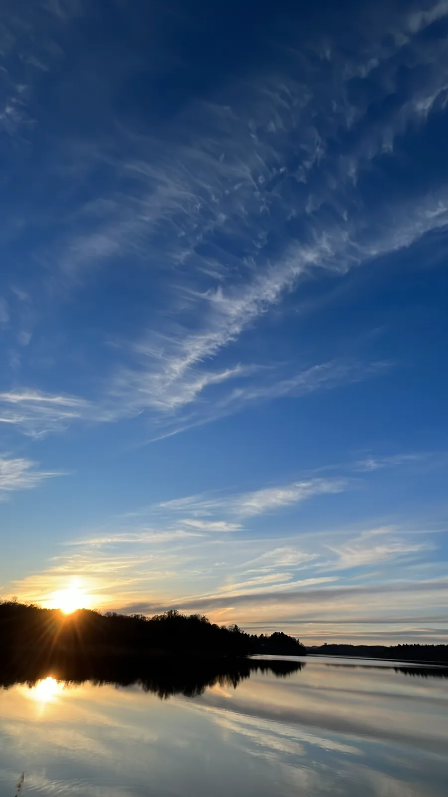 Sunset over a calm lake with wispy cirrus clouds stretching across a deep blue sky